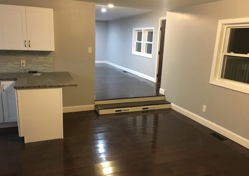 Interior view of a transformed basement with modern kitchen features, including white cabinetry and granite countertop, leading to a well-lit open living area with hardwood flooring and large windows.