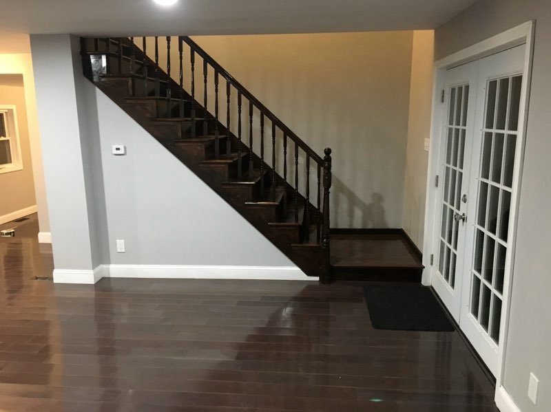 Staircase with dark wooden railing leading to an upper level, featuring polished hardwood flooring and a glass-paneled door, illustrating a transformed basement area into a bright, functional living space.